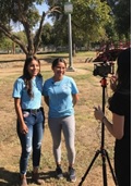 Two young women in light blue shirts in front of a tripod, shown talking to press